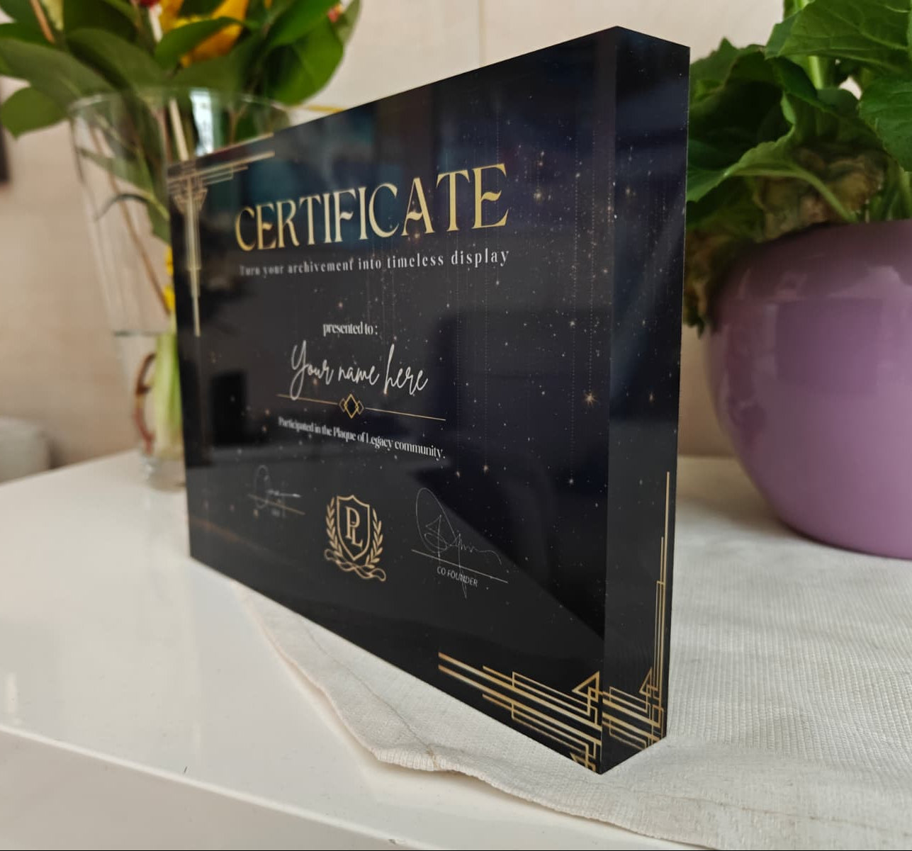 Acrylic certificate holder with gold text on a white surface with plants in the background